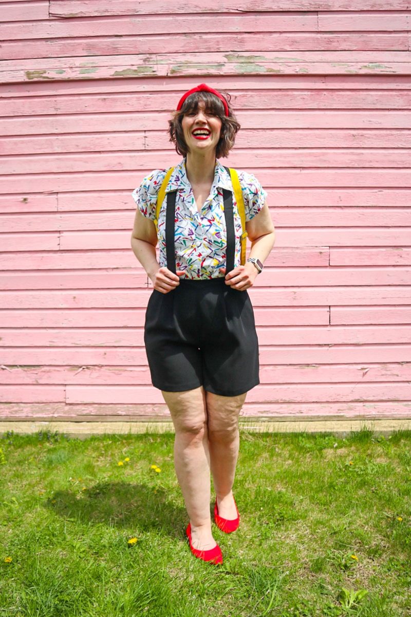 hannah is standing in front of a pink wall. she's wearing red lipstick with matching red headband, a colorful vintage button up, black suspenders, black high waisted shorts, and red ballet flat shoes.