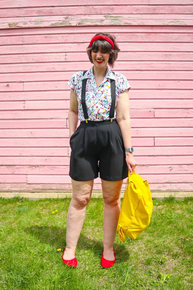 hannah is standing in front of a pink wall. she's wearing red lipstick with matching red headband, a colorful vintage button up, black suspenders, black high waisted shorts, a yellow backpack and red ballet flat shoes.