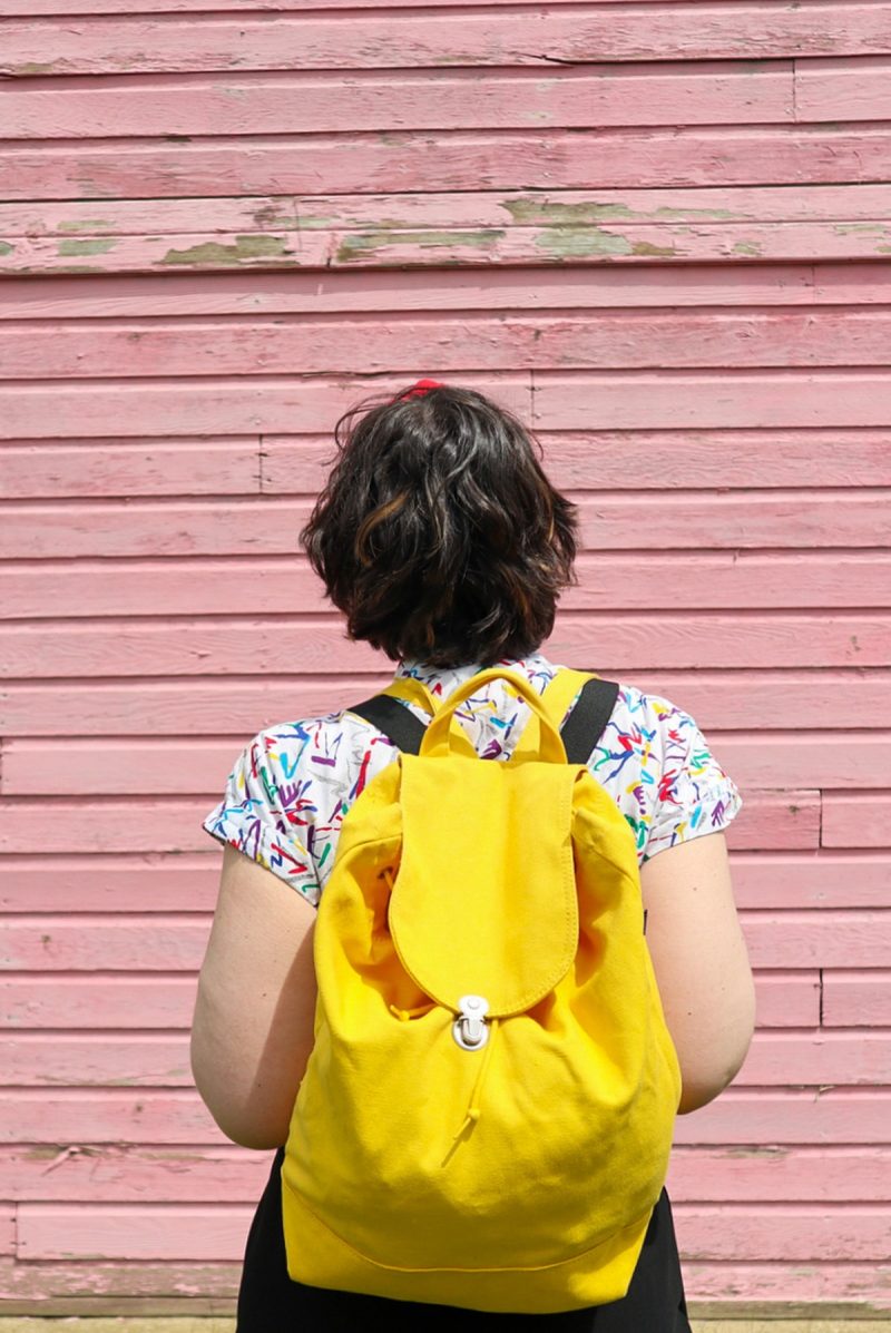 How To Wear Suspenders For Women: 3 Feminine Outfit Ideas | theoutfitrepeater.com you see the back of hannah's head, dark brown curly waves, ands she's looking up at a pink wall. on her back is a big yellow backpack.