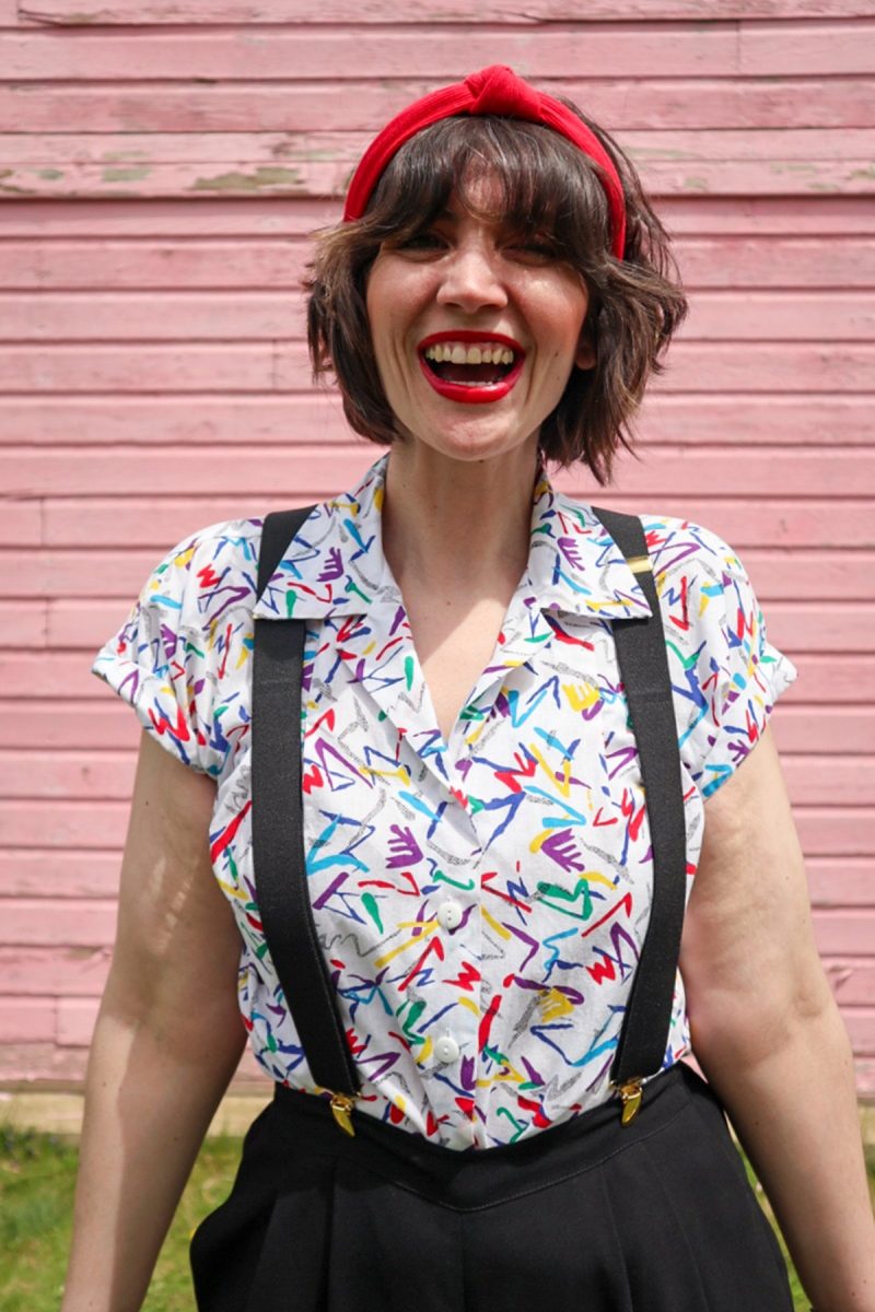 hannah is standing in front of a pink wall. she's wearing red lipstick with matching red headband, a colorful vintage button up, black suspenders, and black high waisted shorts.