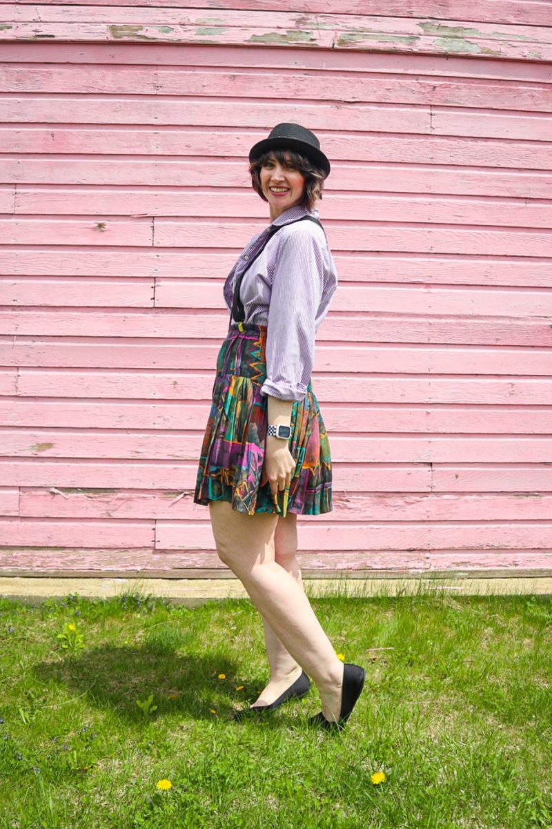 hannah is standing in front of a pink wall. she's wearing a black hat, purple striped button, black suspenders, colorful vintage skirt, and black flat shoes.