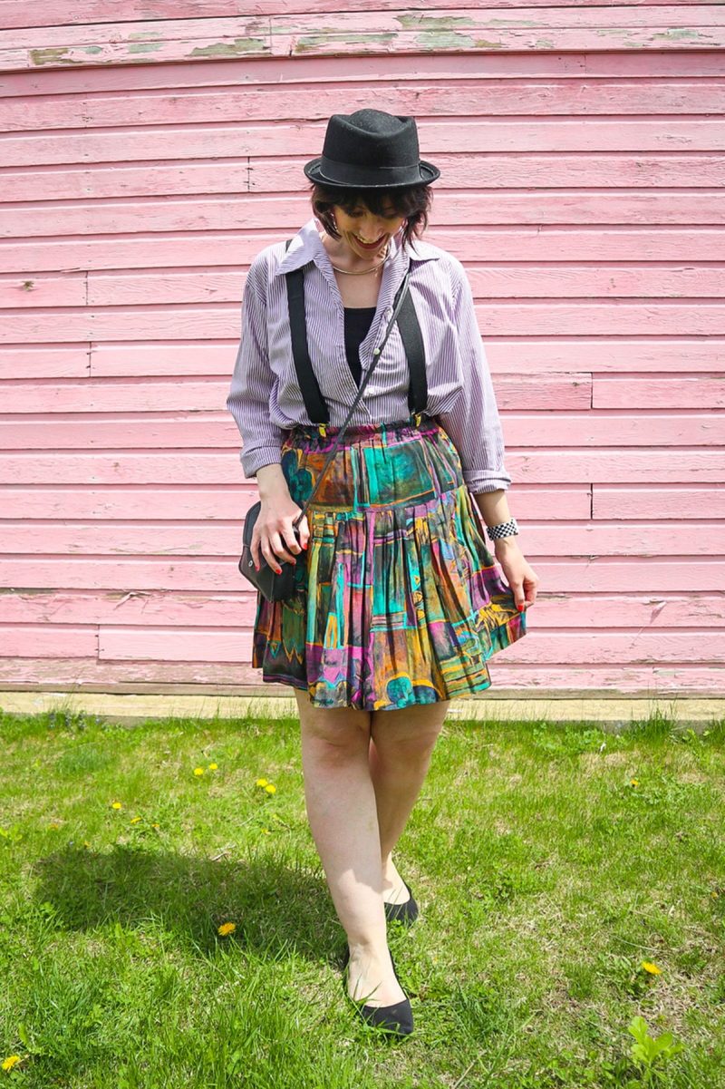 How To Wear Suspenders For Women: 3 Feminine Outfit Ideas | theoutfitrepeater.com hannah is standing in front of a pink wall. she's wearing a black hat, purple striped button, black suspenders, colorful vintage skirt, and black flat shoes.