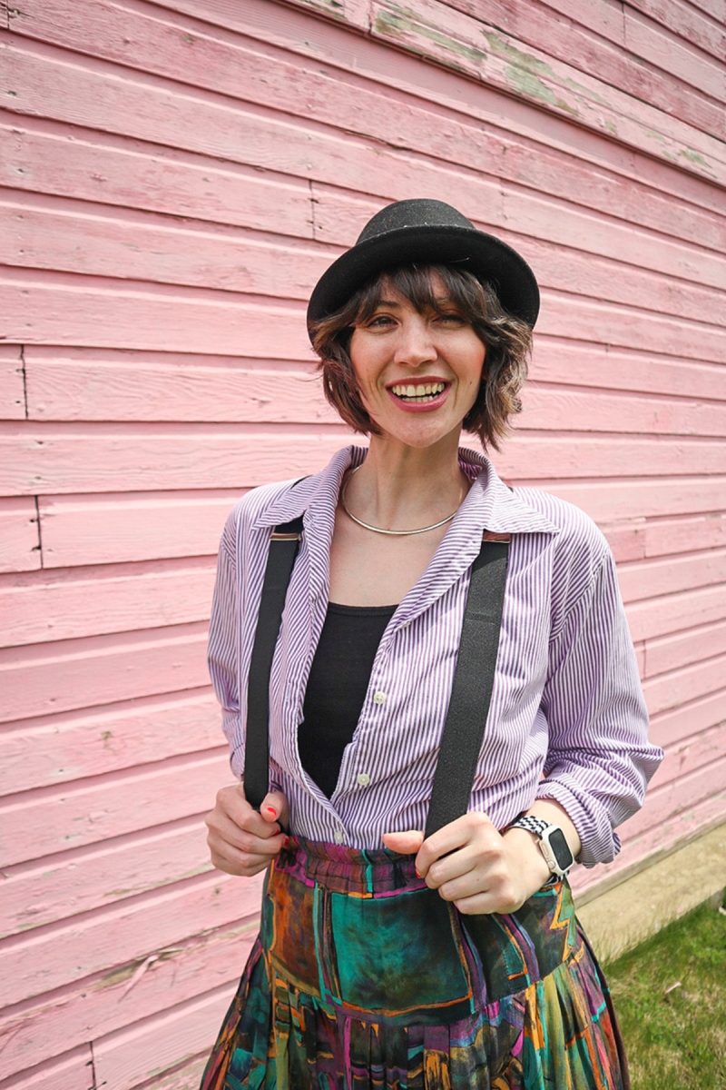 hannah is standing in front of a pink wall. she's wearing a black hat, purple striped button, black suspenders, and a colorful vintage skirt