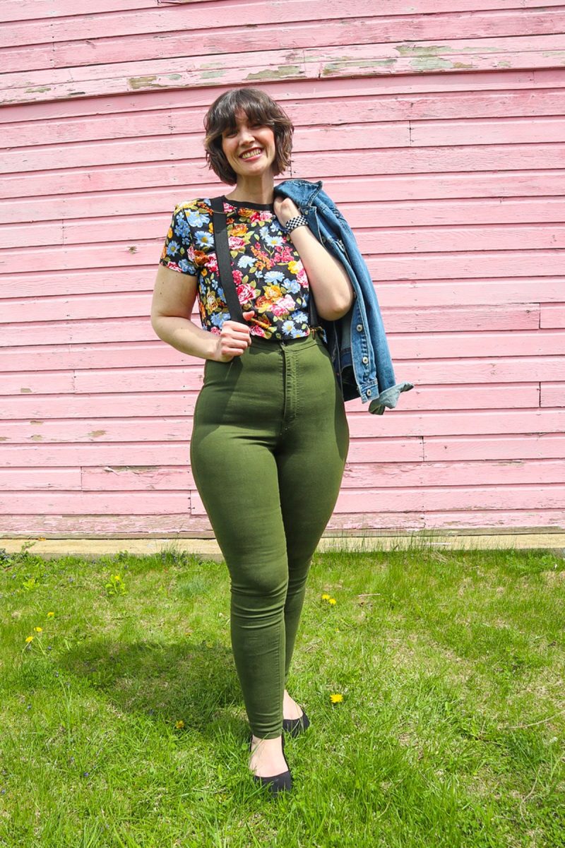 How To Wear Suspenders For Women: 3 Feminine Outfit Ideas | theoutfitrepeater.com hannah is standing in front of a pink wall. she's wearing a floral crop top, denim jacket, black suspenders, olive green skinny pants, and a black flat shoes.