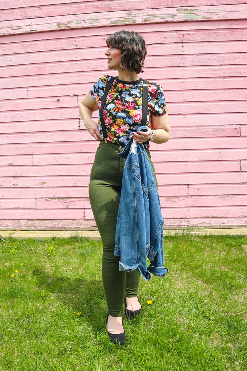 hannah is standing in front of a pink wall. she's wearing a floral crop top, denim jacket, black suspenders, olive green skinny pants, and a black flat shoes.