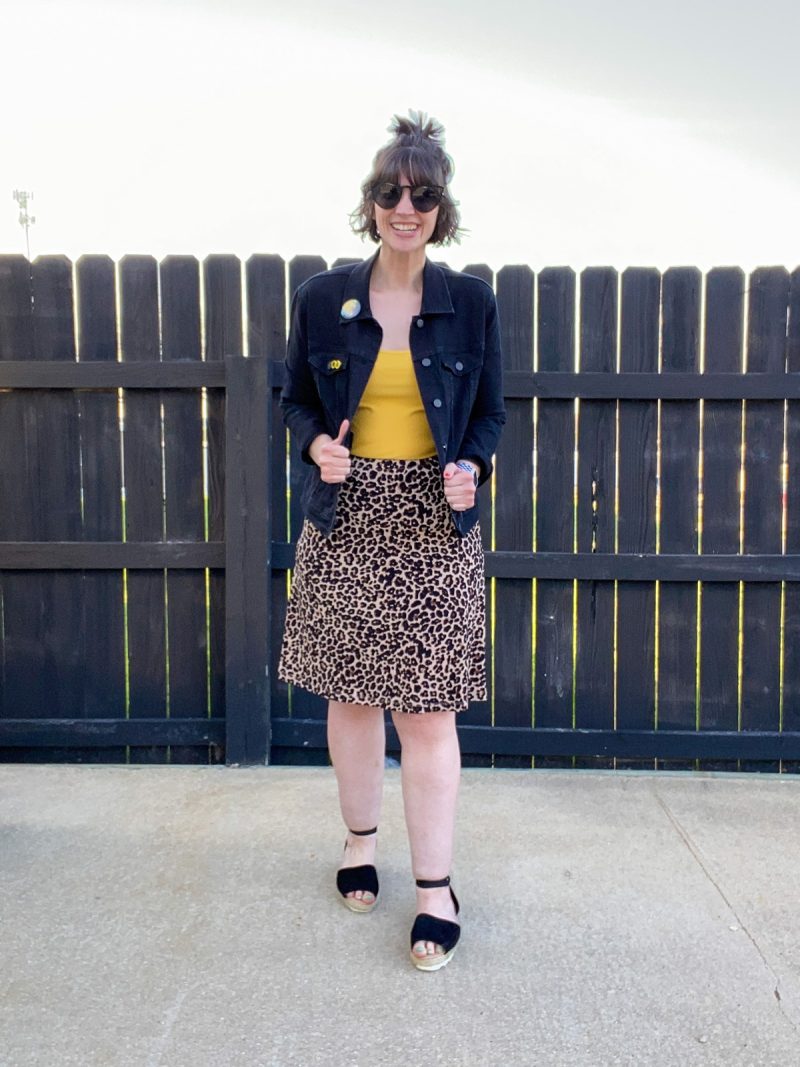 Wearing All Black Is Boring, Here's How To Make It Better | www.theoutfitrepeater.com hannah rupp is a white female with short brown hair. she's wearing dark sunglasses, a black denim jacket, yellow tank top, leopard print skirt, and black sandals.