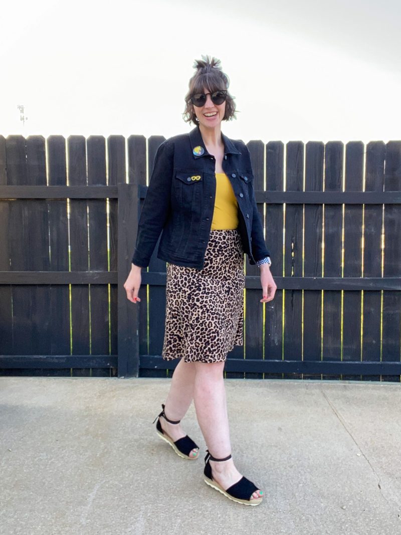Wearing All Black Is Boring, Here's How To Make It Better | www.theoutfitrepeater.com hannah rupp is a white female with short brown hair. she's wearing dark sunglasses, a black denim jacket, yellow tank top, leopard print skirt, and black sandals.
