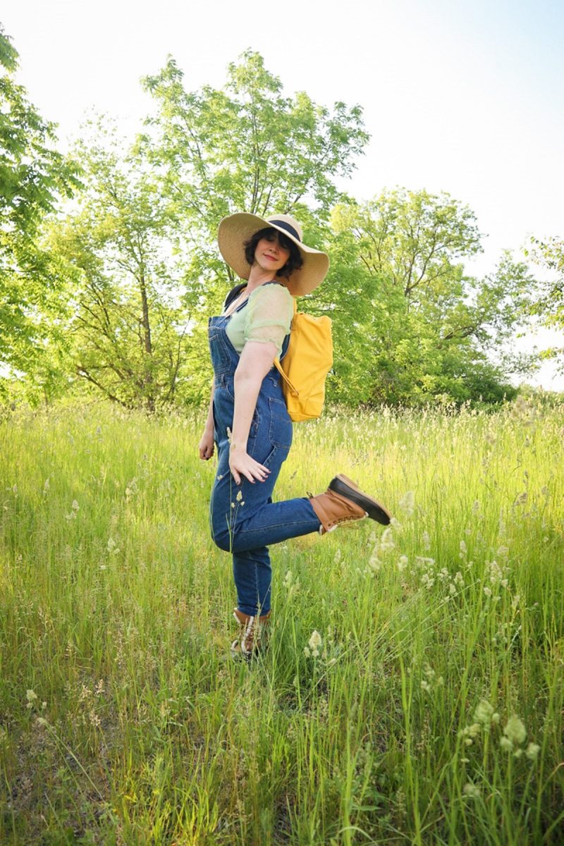 This Outfit Will Convince You To Wear Those Fancy Clothes You've Been Saving | www.theoutfitrepeater.com hannah rupp is in a bright, green field. she's wearing a mint green blouse, floppy hat, denim overalls, snow boots, and a yellow backpack.