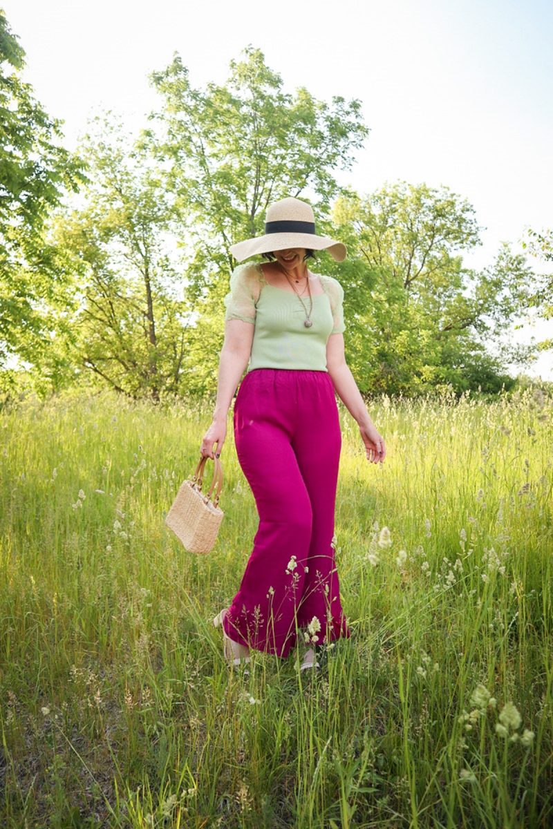 hannah rupp is in a bright, green field. she's wearing a mint green blouse, floppy hat, magenta palazzo pants, straw bag, and gold jewelry.