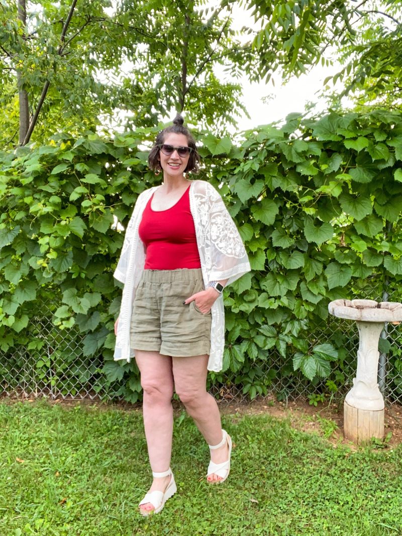 hannah is standing in a yard surrounded by greenery. she's wearing a red tank top, white kimono, olive green shorts, and white platform sandals.
