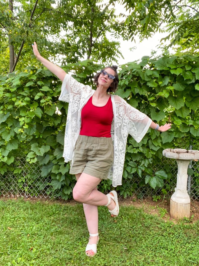 hannah is standing in a yard surrounded by greenery. she's wearing a red tank top, white kimono, olive green shorts, and white platform sandals.