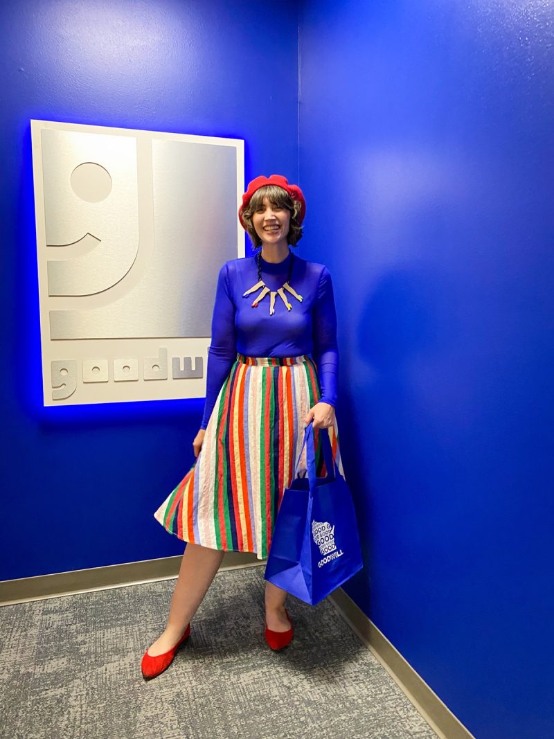 hannah rupp wears a bright blue shirt, rainbow skirt, and standing in front of the goodwill logo