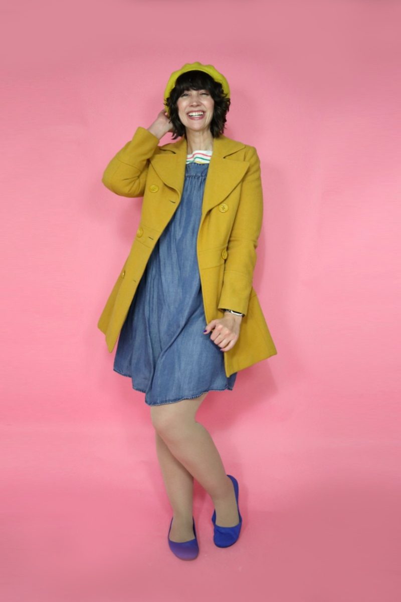 hannah is standing in front of a pink background, smiling, wearing a denim dress, yellow pea coat, yellow beret, and cobalt blue flats.