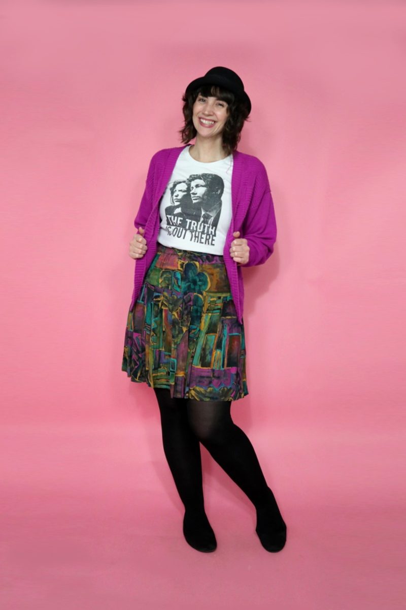 hannah has curly hair worn under a black pork pie hat. she's smiling while wearing an x-files graphic tee, purple cardigan, colorful skirt, black tights, and black ballet flats