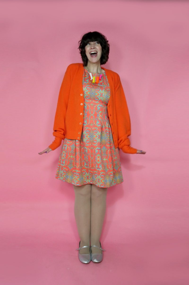 hannah is wearing an orange patterned dress, an orange cardigan, skin tone colored tights, and silver mary jane flats.