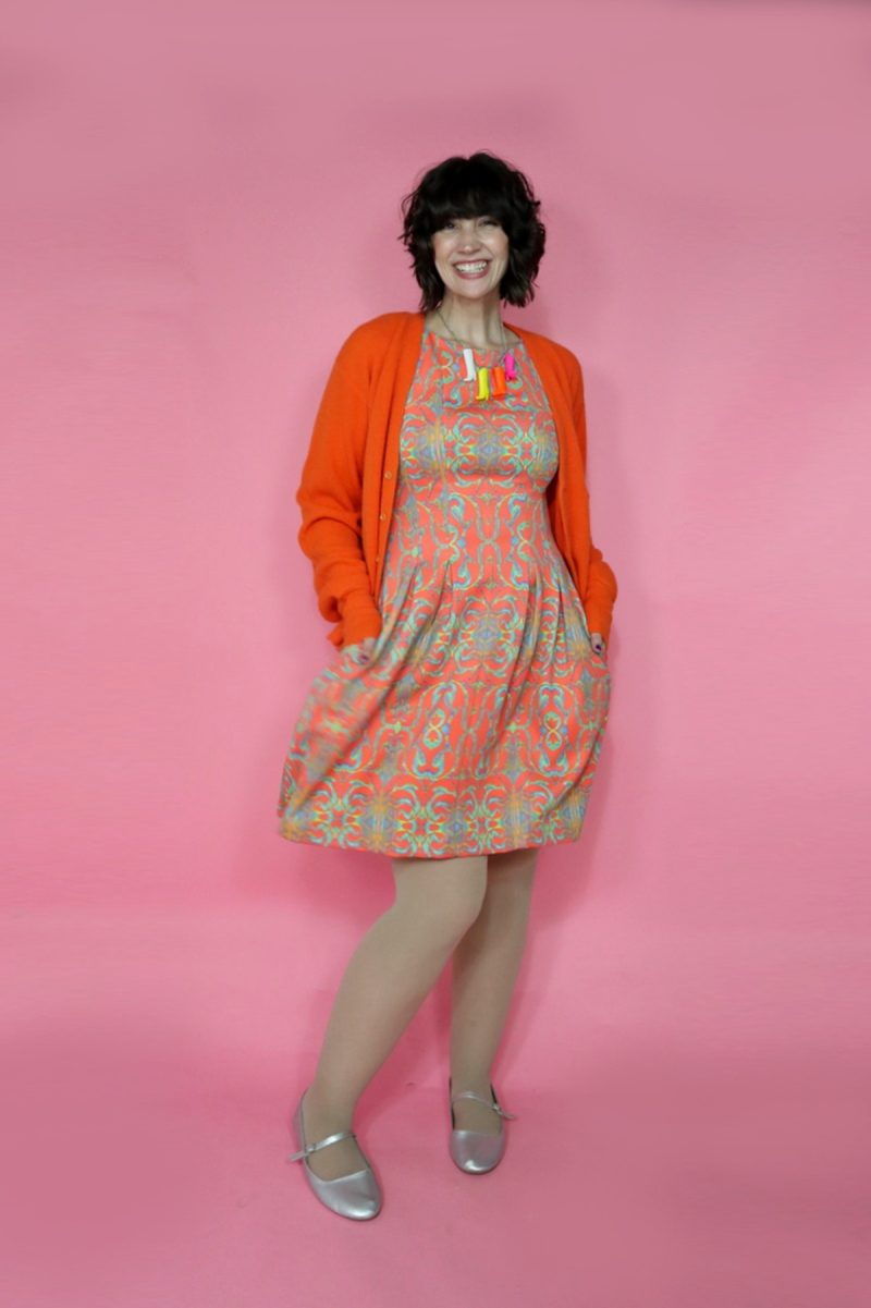 hannah is wearing an orange patterned dress, an orange cardigan, skin tone colored tights, and silver mary jane flats.