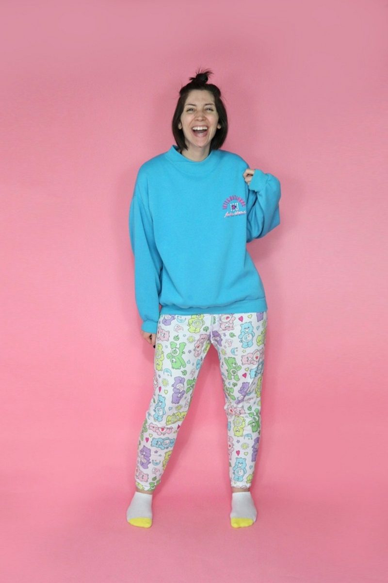 hannah is a white female with short brown hair. she's smiling in front of a pink background. she's wearing an oversized blue sweatshirt, care bear pajama pants, and pink fuzzy slippers