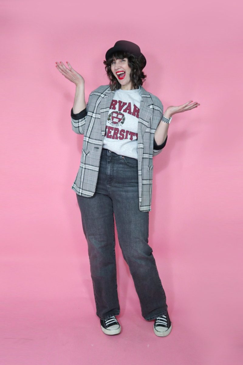 hannah is a white woman with curly brown hair. she's standing in front of a pink background. she's wearing a gray harvard sweatshirt, plaid blazer, black hat, gray wide leg jeans, and black converse style sneakers.