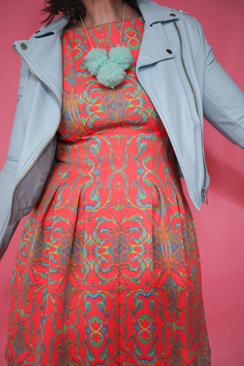 hannah is wearing an orange patterned dress, light blue moto jacket, and teal pom pom necklace