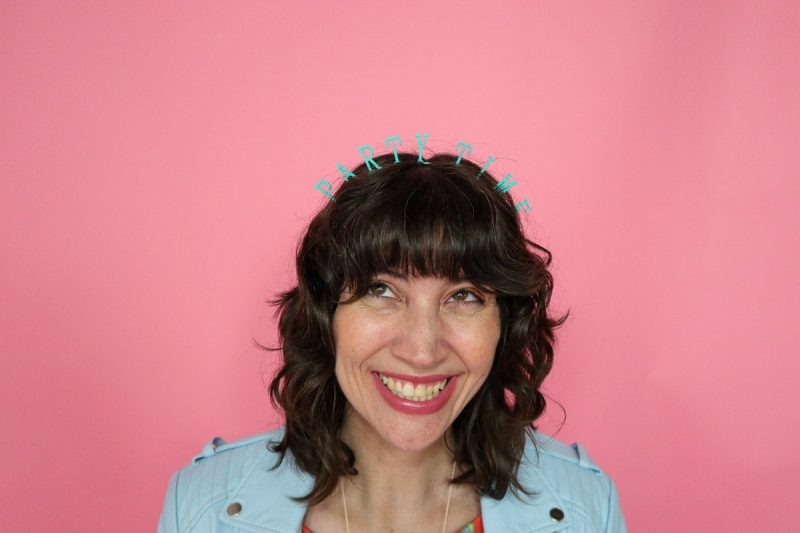 hannah is a light skin woman with short dark curly hair. she's smiling and wearing a headband that says "party time"