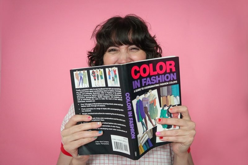 Budget-Friendly Personal Color Analysis: Affordable Ways to Find Your Palette | www.theoutfitrepeater.com white female with brown curly hair, pink paper background, holding a vintage book titled color in fashion