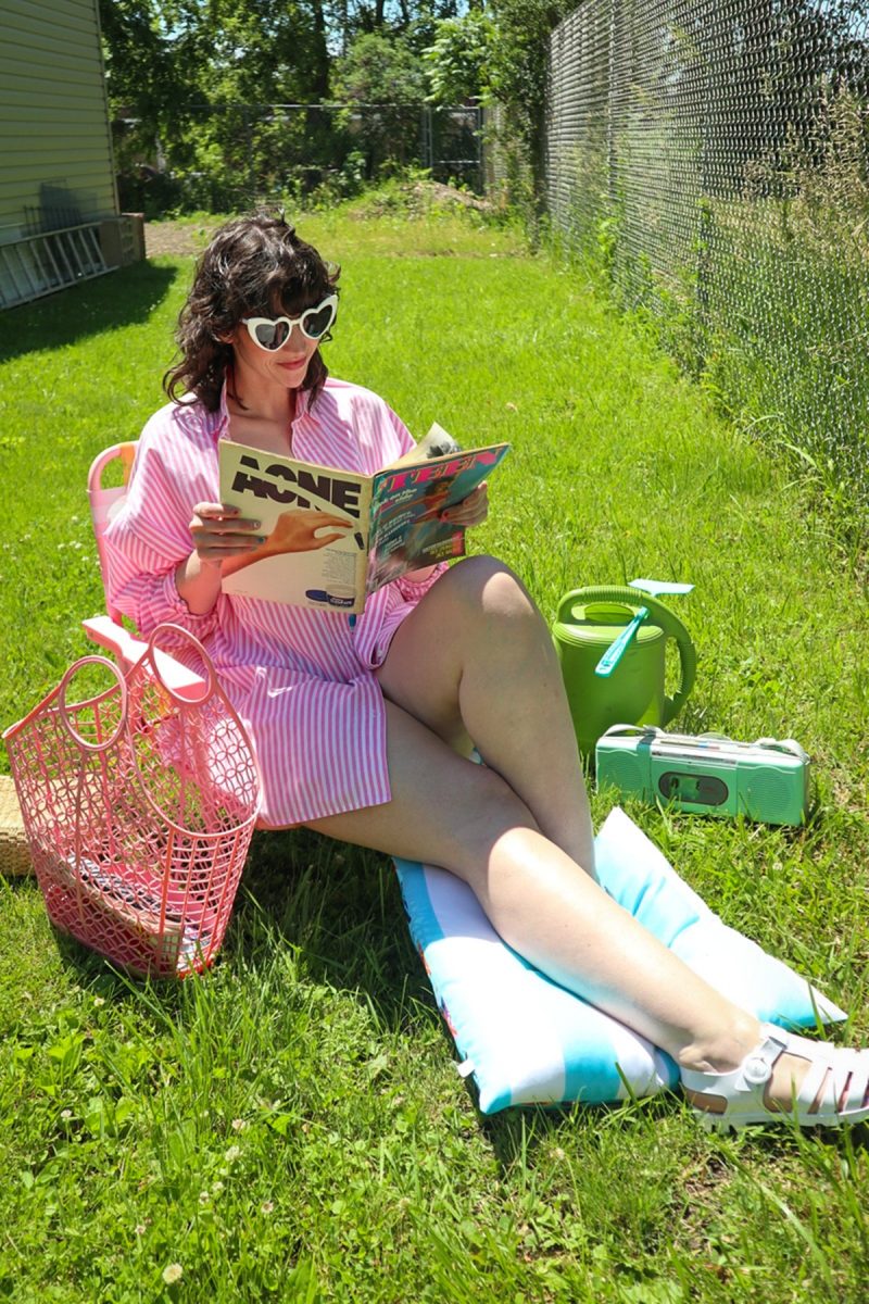 The Dream Thrifted Swimsuit I Scored for under $10 - And How I Styled It! | www.theoutfitrepeater.com a smiling white female with brown curly hair is sitting outside on a sunny day. she's wearing an oversized pink and white button up and reading a magazine.