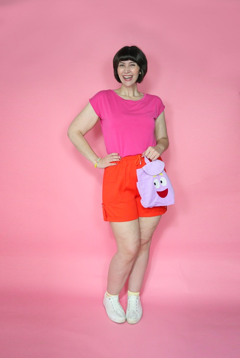 a dora the explorer halloween costume portrayed by a white millennial women with short brown bob hair, pink t-shirt, orange shorts, and purple backpack