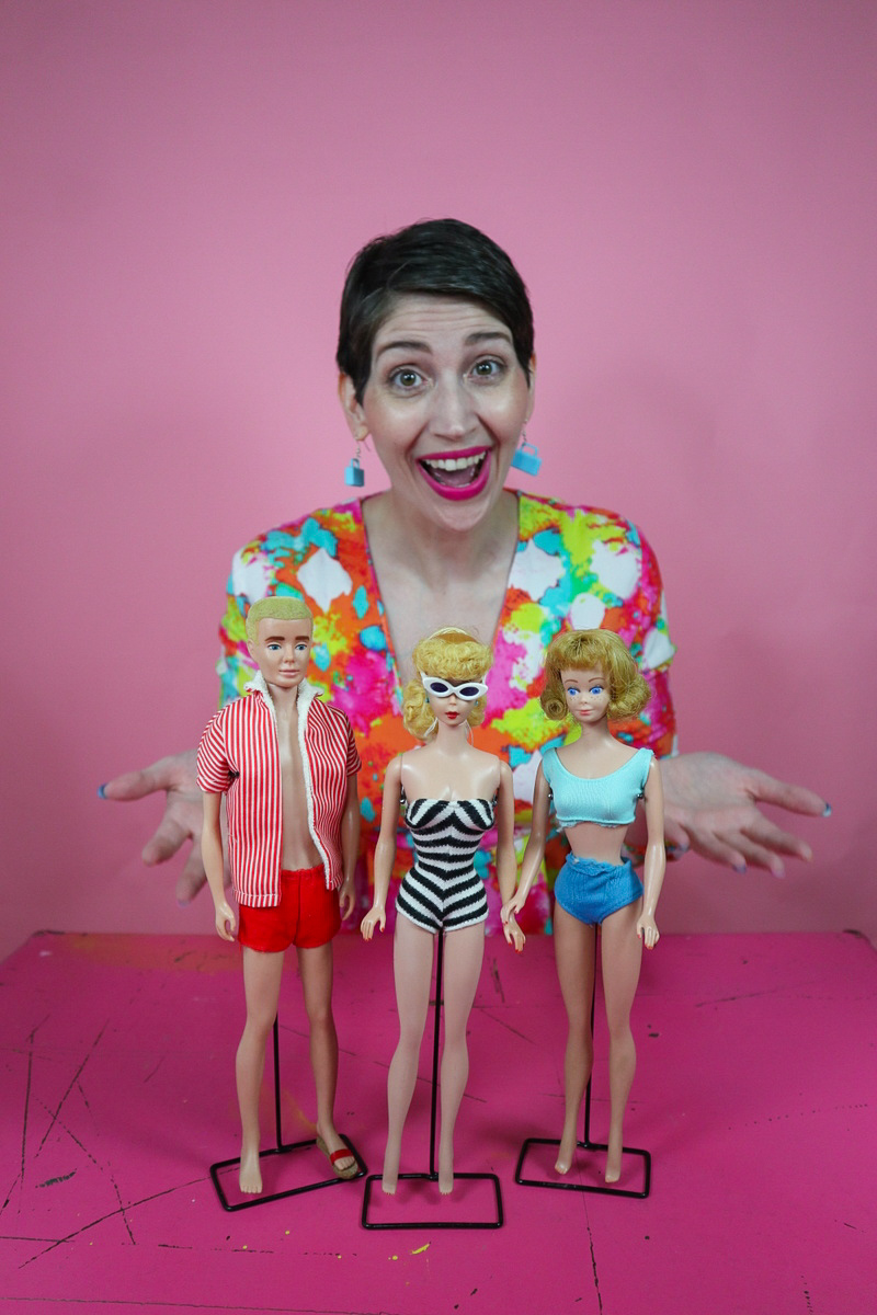a young white female with a brown pixie cut and colorful dress is smiling. in front of her is a display of three vintage Barbie dolls. 