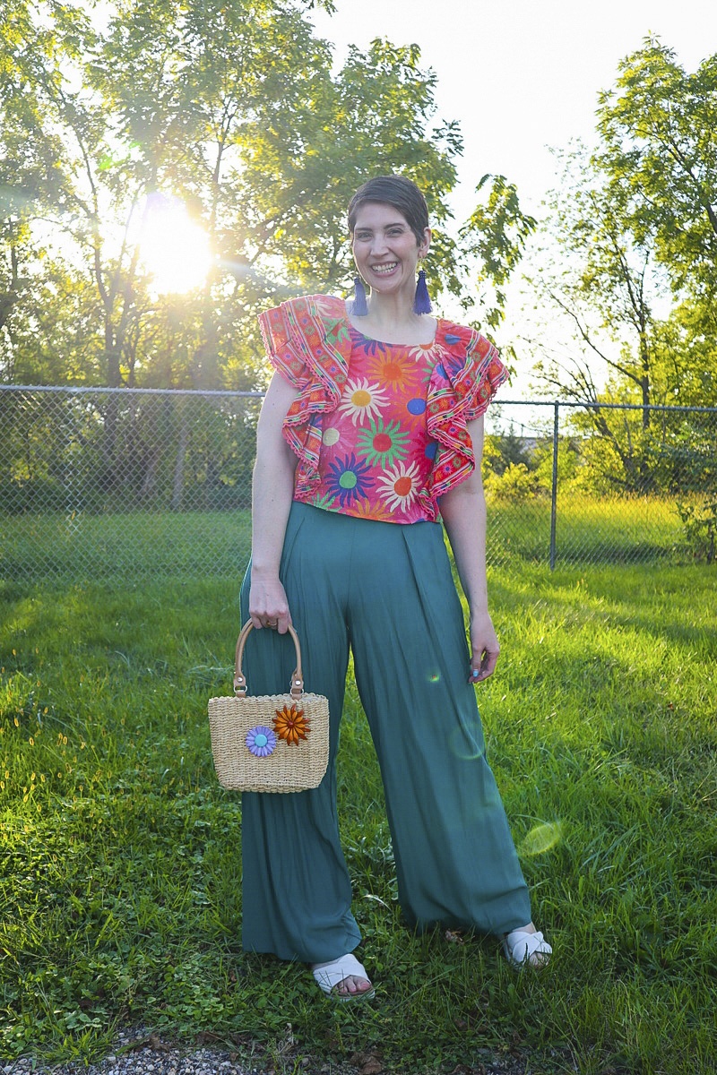What I Thrifted vs. How I Styled It | www.theoutfitrepeater.com a white female with a short brown pixie cut is smiling outdoors while wearing a bright patterned top, teal palazzo pants, and a thrifted basket weaved purse