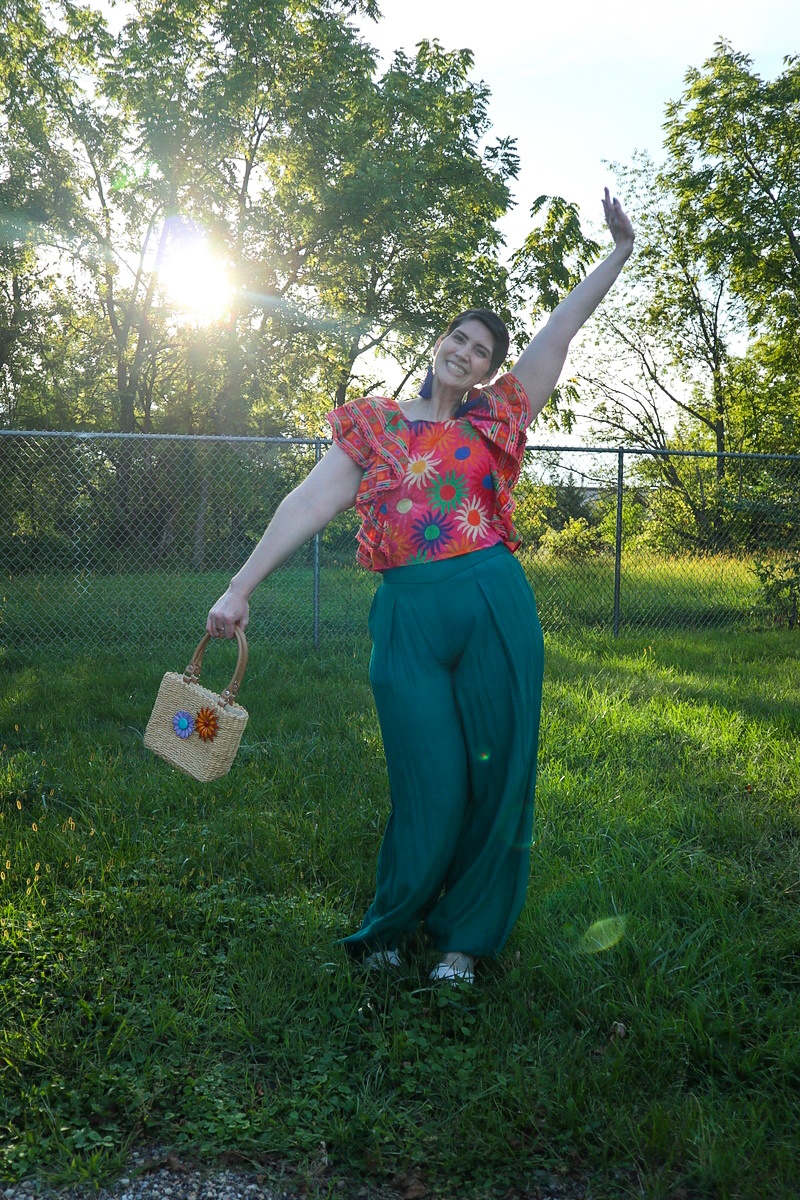 What I Thrifted vs. How I Styled It | www.theoutfitrepeater.com a white female with a short brown pixie cut is smiling outdoors while wearing a bright patterned top, teal palazzo pants, and a thrifted basket weaved purse