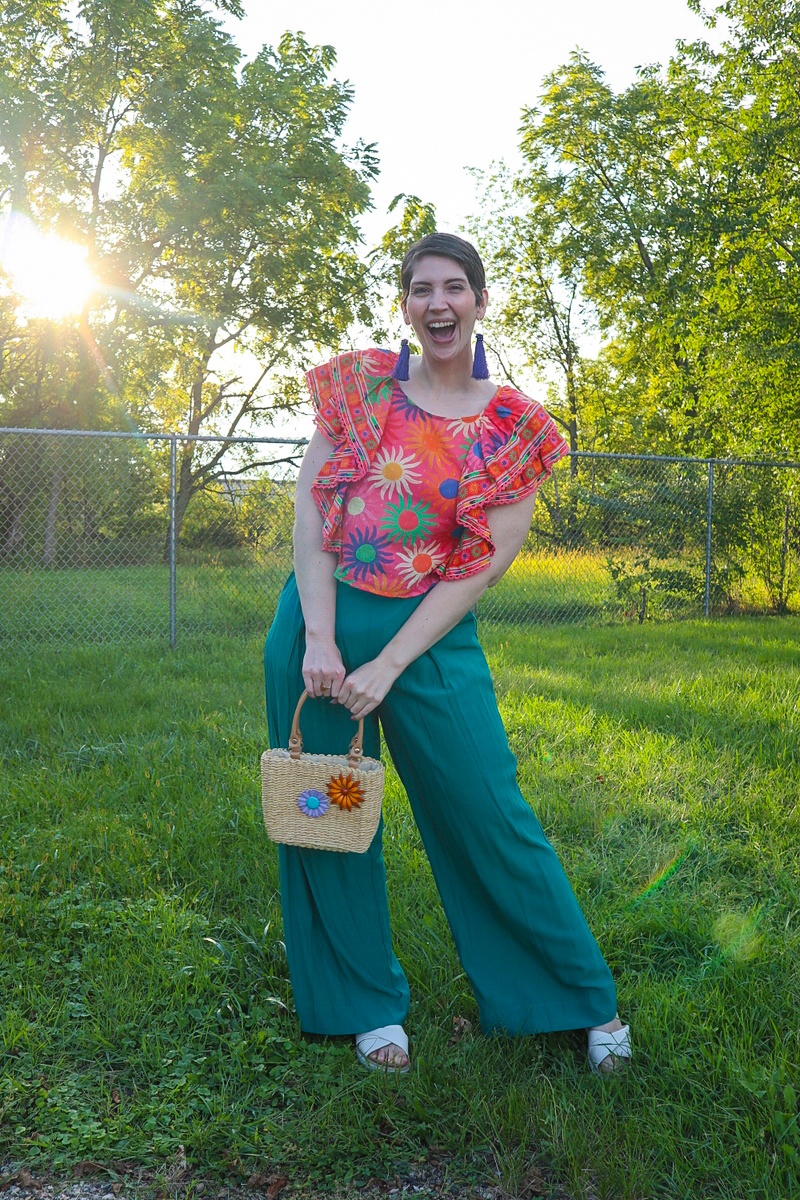 a white female with a short brown pixie cut is smiling outdoors while wearing a bright patterned top, teal palazzo pants, and a thrifted basket weaved purse