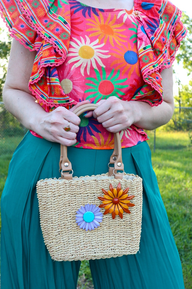 What I Thrifted vs. How I Styled It | www.theoutfitrepeater.com a white female is outdoors, holding a basket weaved handbag with vintage floral brooches on it. she's wearing a bright patterned top and teal pants.