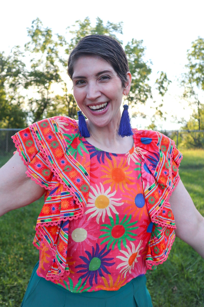 What I Thrifted vs. How I Styled It | www.theoutfitrepeater.com a white female with a short brown pixie cut is smiling outdoors while wearing a bright patterned top and blue tassel earrings